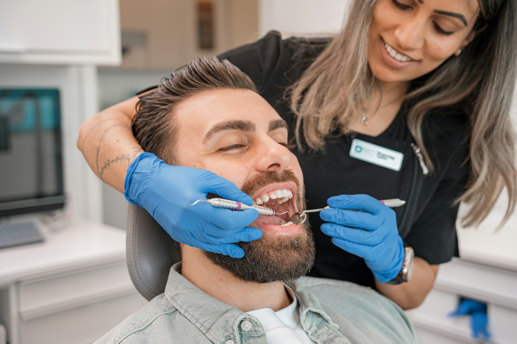 A dentist smiling as she checks a relaxed patient's teeth in a bright treatment room.